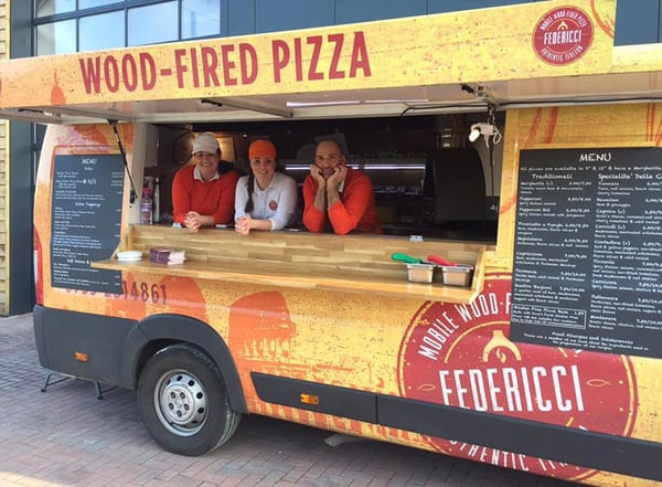 Two women and a man looking out from the hatch of a wood-fired pizza food truck